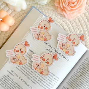 Puede incluir: Cuatro pegatinas con un pollito de dibujos animados llorando leyendo un libro, con el texto "crying over fictional characters". Las pegatinas son de color amarillo claro con detalles en naranja y un pequeño lazo. Las pegatinas están colocadas sobre un libro abierto.