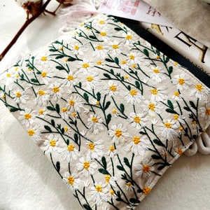 May include: A beige fabric pouch with a white daisy floral embroidery pattern. The pouch is made of a soft, natural fabric and features a delicate floral design.