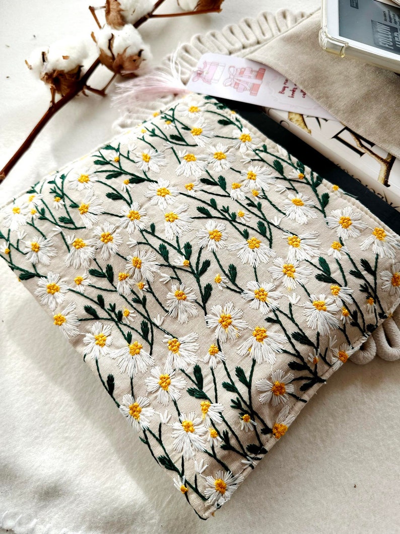 May include: A beige fabric pouch with a white daisy floral embroidery pattern. The pouch is made of a soft, natural fabric and features a delicate floral design.