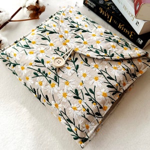 May include: A beige fabric book cover with a white daisy floral embroidery pattern. The cover has a flap with a button closure and a green drawstring.
