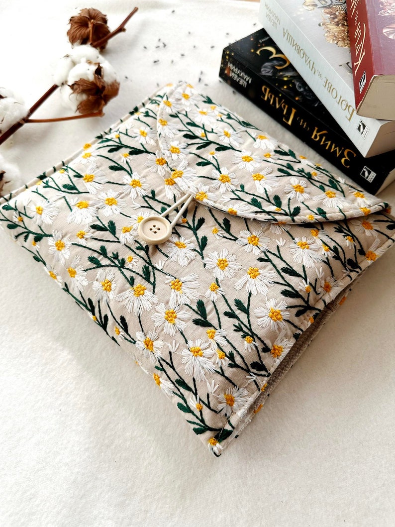 May include: A beige fabric book cover with a white daisy floral embroidery pattern. The cover has a flap with a button closure and a green drawstring.