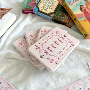 May include: A collection of books and accessories on a white surface. The books include titles like "Ride the Wave" and "The Cinnamon Bun Book Store." Pink coasters with the text "Pretty Girls Read Books" are also visible, along with bookmarks.