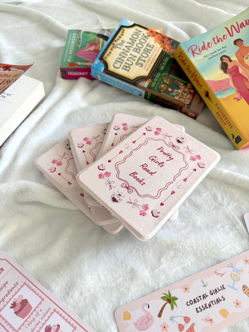 May include: A collection of books and accessories on a white surface. The books include titles like "Ride the Wave" and "The Cinnamon Bun Book Store." Pink coasters with the text "Pretty Girls Read Books" are also visible, along with bookmarks.