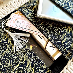 May include: A black fabric book sleeve with a gold cloud pattern. The sleeve has a zippered pocket on the top and a bookmark with the text "Daydream Club - Busy obsessing over morally gray characters."