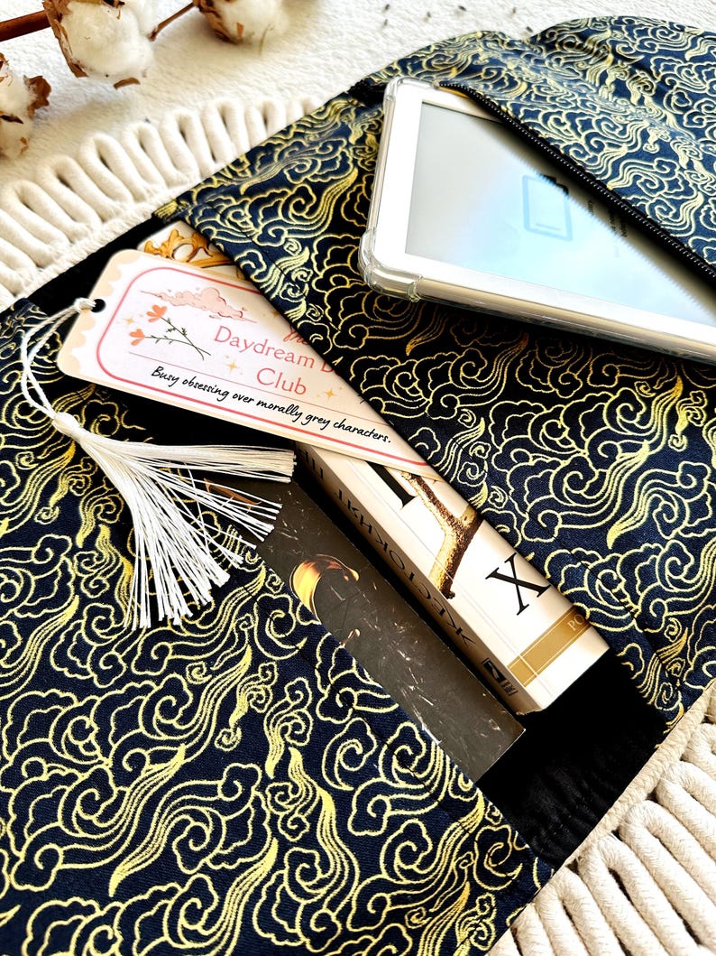 May include: A black fabric book sleeve with a gold cloud pattern. The sleeve has a zippered pocket on the top and a bookmark with the text "Daydream Club - Busy obsessing over morally gray characters."