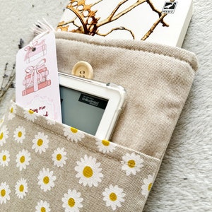 May include: A beige canvas book sleeve with a white daisy pattern. The sleeve has a pocket on the front for a book or e-reader. A white e-reader is in the pocket, and a book with a gold design is in the top pocket. The book has the text "KINDLE UNLIMITED" on the cover.