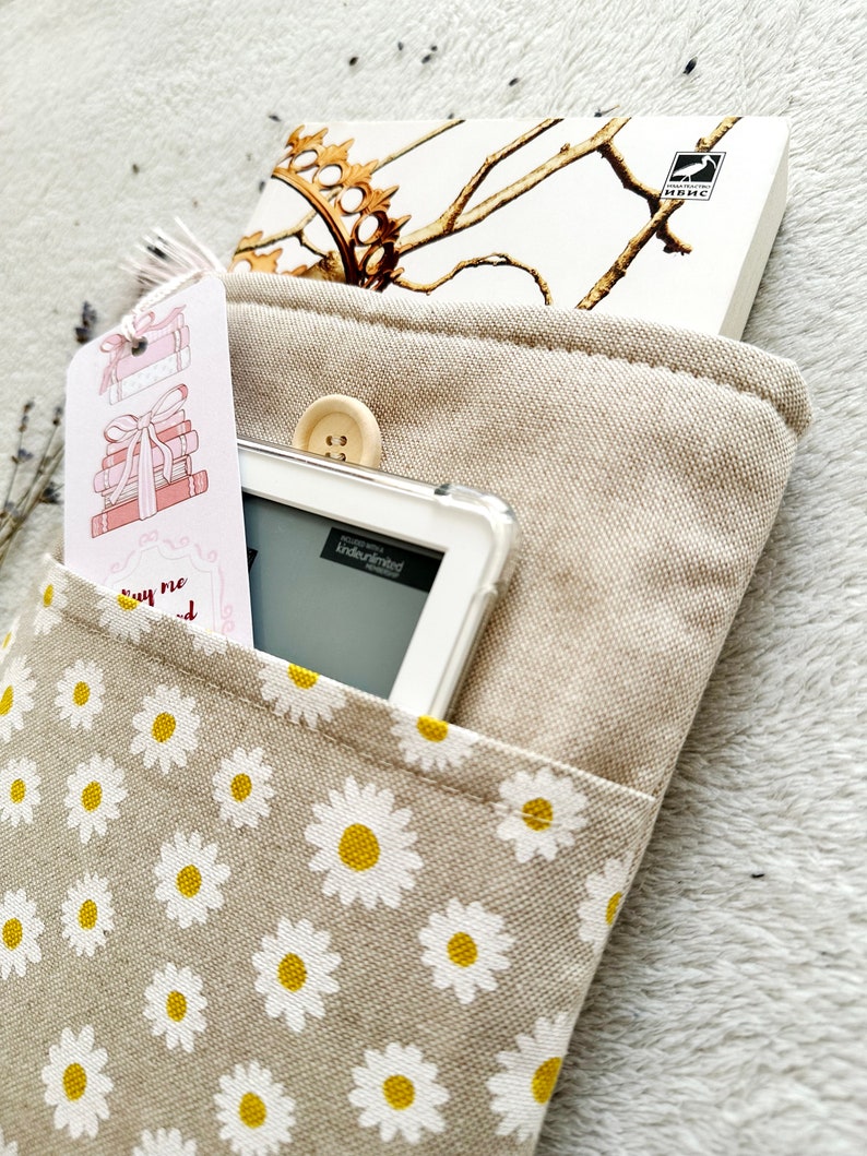 May include: A beige canvas book sleeve with a white daisy pattern. The sleeve has a pocket on the front for a book or e-reader. A white e-reader is in the pocket, and a book with a gold design is in the top pocket. The book has the text "KINDLE UNLIMITED" on the cover.