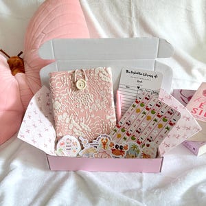 Puede incluir: Una caja de regalo rosa llena de una cubierta de libro, pegatinas, un bloc de notas y papel estampado. La cubierta del libro es rosa claro con un diseño floral y un cierre de botón. Las pegatinas presentan ilustraciones con temática de playa. La caja está sobre una superficie blanca.