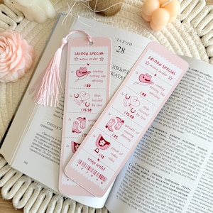 Op de afbeelding: Twee roze bladwijzers met kwastjes liggen op een open boek. De bladwijzers hebben een saloon-thema met cowboyhoedillustraties en tekst, waaronder "Saloon Special" en "Stay Wild."