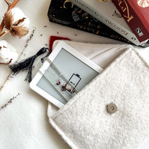 Może przedstawiać: Biała torebka z filcu z zamkiem błyskawicznym i srebrnym guzikiem. Wewnątrz torebki znajduje się biały czytnik e-book&oacute;w z ikoną baterii na ekranie.