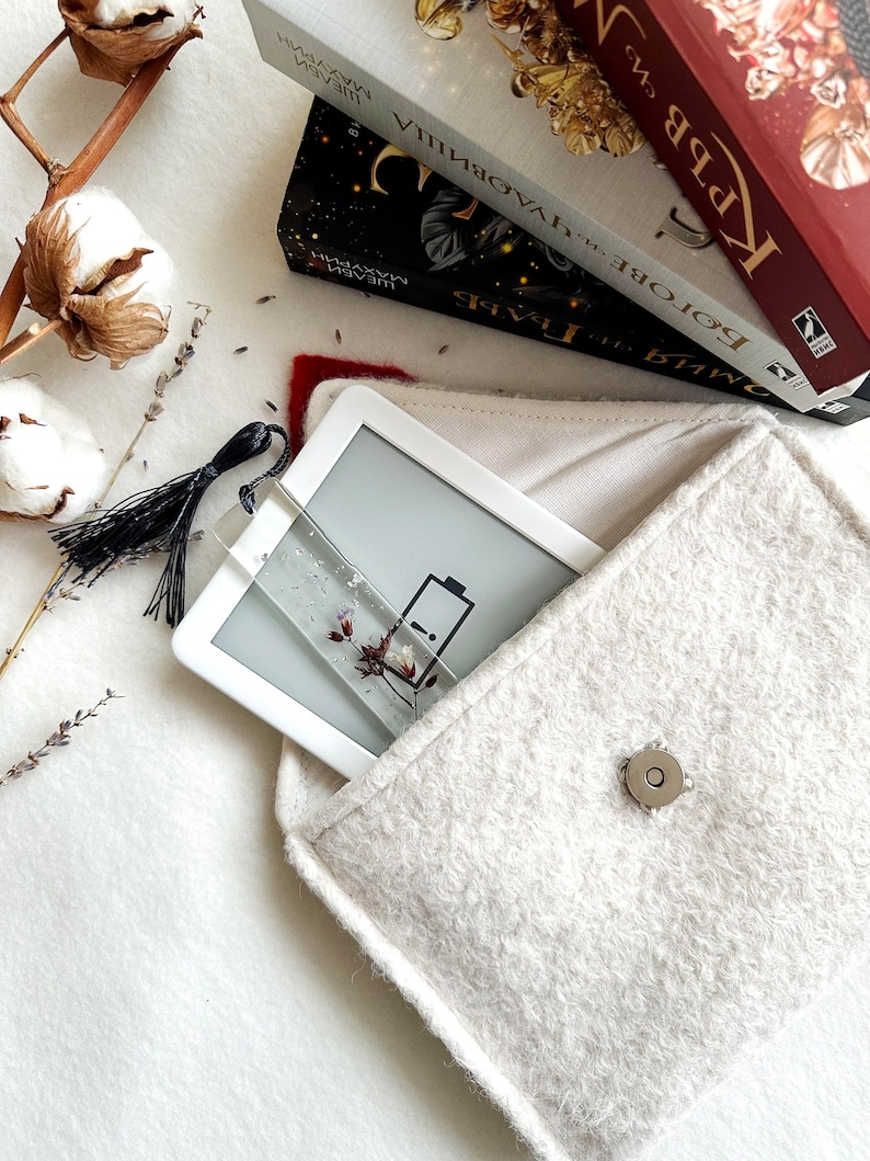 Może przedstawiać: Biała torebka z filcu z zamkiem błyskawicznym i srebrnym guzikiem. Wewnątrz torebki znajduje się biały czytnik e-book&oacute;w z ikoną baterii na ekranie.