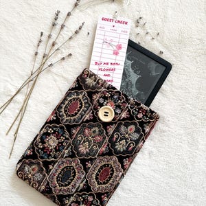 May include: A black fabric sleeve with an intricate, multi-colored floral and geometric embroidery pattern, secured by a single light-colored wooden button. A white bookmark shaped like a guest check with the text "GUEST CHECK" and "BUY ME BOTH FLOWERS AND BOOKS" is tucked inside.