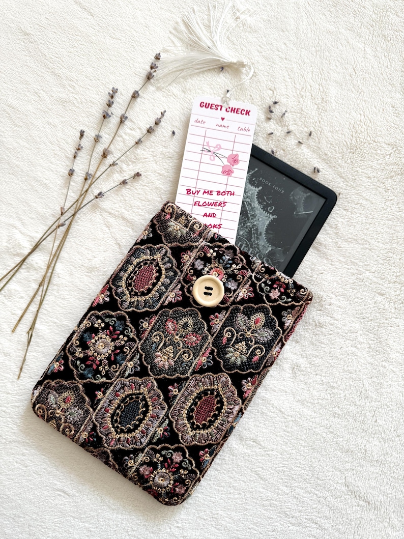 May include: A black fabric sleeve with an intricate, multi-colored floral and geometric embroidery pattern, secured by a single light-colored wooden button. A white bookmark shaped like a guest check with the text "GUEST CHECK" and "BUY ME BOTH FLOWERS AND BOOKS" is tucked inside.