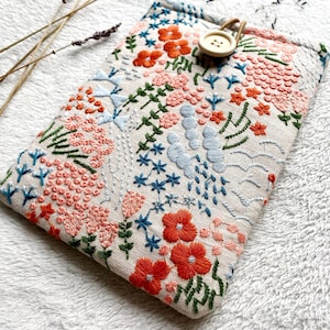 May include: A beige linen pouch with a floral embroidery design. The embroidery features pink, orange, blue, and green flowers and leaves. A wooden button is attached to the pouch.