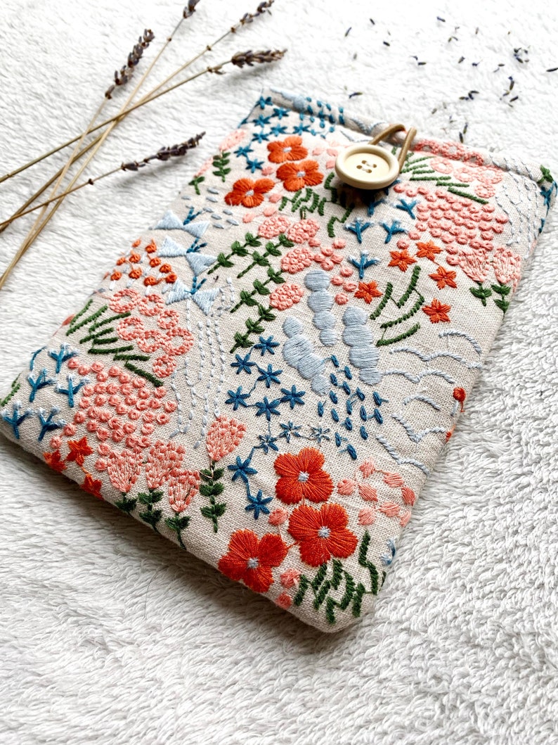 May include: A beige linen pouch with a floral embroidery design. The embroidery features pink, orange, blue, and green flowers and leaves. A wooden button is attached to the pouch.