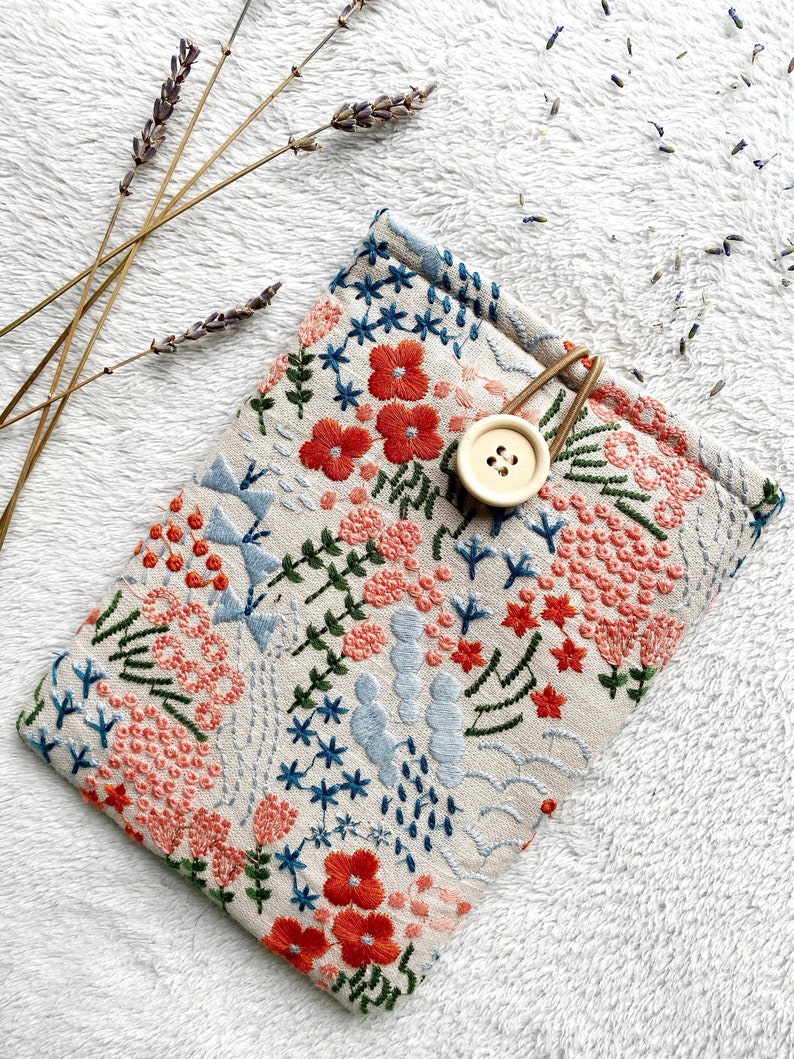 May include: A beige fabric pouch with a floral embroidery design in shades of blue, green, and orange. The pouch has a wooden button closure and a gold metal clasp.