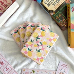 May include: A stack of pink phone cases with a lemon and floral pattern. Books and bookmarks are arranged around the cases. The bookmarks feature text and illustrations. One bookmark reads "Coastal Girlie Essentials".