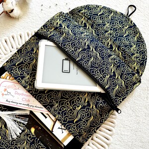 May include: A black and gold fabric book sleeve with a zipper closure. The sleeve has a cloud pattern and is open to reveal a white e-reader and a book. The text on the book reads "Daydream Club: Busy observing our morally grey characters."