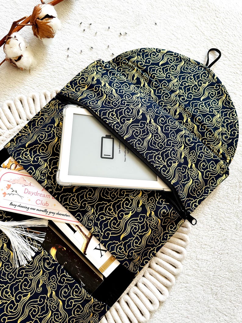 May include: A black and gold fabric book sleeve with a zipper closure. The sleeve has a cloud pattern and is open to reveal a white e-reader and a book. The text on the book reads "Daydream Club: Busy observing our morally grey characters."