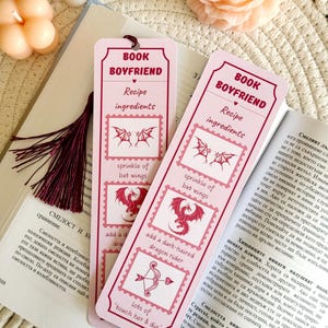 May include: Two pink bookmarks with the text "Book Boyfriend" and "Recipe Ingredients" on them. The bookmarks feature illustrations of bats, dragons, and an arrow with hearts. The text "sprinkle of bat wings", "add a dark-haired dragon rider", and "lots of touch her & die" are also on the bookmarks.