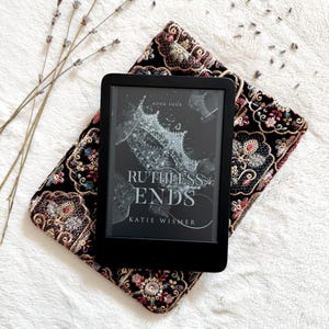 May include: A black e-reader displaying the book cover for Ruthless Ends, Book Four by Katie Wismer, resting on a black velvet pouch featuring intricate, multi-colored floral embroidery with gold and pink thread accents.