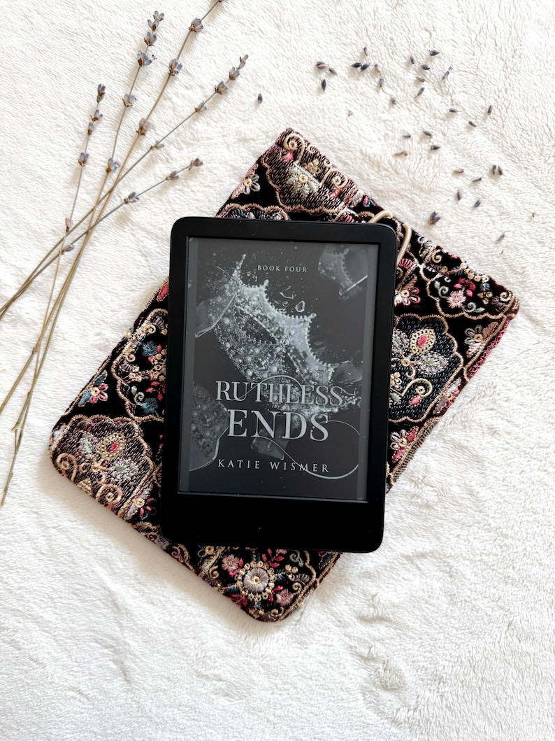 May include: A black e-reader displaying the book cover for Ruthless Ends, Book Four by Katie Wismer, resting on a black velvet pouch featuring intricate, multi-colored floral embroidery with gold and pink thread accents.