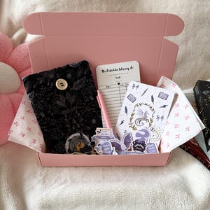 Puede incluir: Una caja rosa llena de artículos temáticos de libros. En el interior hay una funda de libro negra, pegatinas, un bloc de notas y un bolígrafo rosa. La funda del libro tiene un cierre de botón. El bloc de notas tiene una ilustración de un dragón y el texto "The Forbidden Library of: Goals".