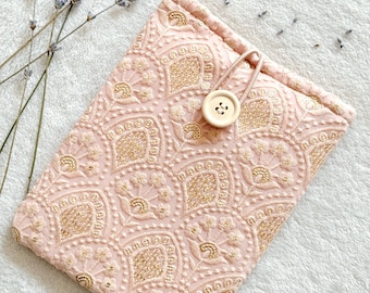 Puede incluir: Una bolsa de tela rosa con un patrón bordado en oro y un cierre de botón de madera. La bolsa está sobre una superficie blanca y peluda.