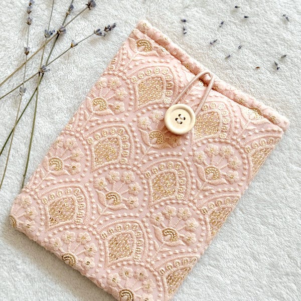 Könnte beinhalten: Eine rosa Stofftasche mit einem goldenen Stickmuster und einem Verschluss aus Holz. Die Tasche liegt auf einer weißen, flauschigen Oberfläche.