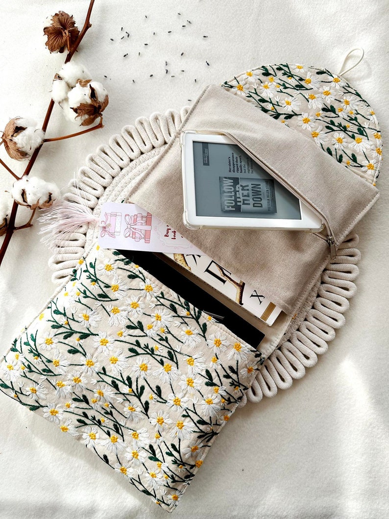 May include: A white canvas pouch with a zipper closure and a daisy floral embroidery design. The pouch is open and a white e-reader is inside. The e-reader screen displays the text "Follow Her Down" and the author's name.