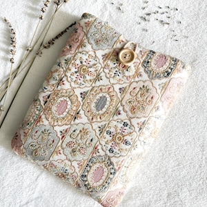 May include: A rectangular fabric pouch featuring a repeating pattern of ornate, scalloped-edge medallions in shades of pink, gold, blue, and cream. The pouch is secured with a light-coloured elastic loop and a single, round, light-brown wooden button.