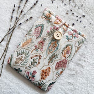 Könnte beinhalten: Cremefarbene Buchhülle mit einem gestickten Blumenmuster in Rosa-, Türkis- und Orangetönen. Die Hülle wird mit einem Holzknopf und einer Schlaufe verschlossen. Getrocknete Lavendelzweige sind in der Nähe der Hülle platziert.