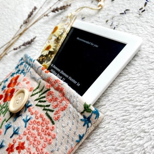 May include: A white e-reader with a floral bookmark in a beige fabric case with embroidered flowers. The e-reader screen displays the text "Recommended for you" and the title of a book, "A Demons Demon Hunter in Hiding Book 1".
