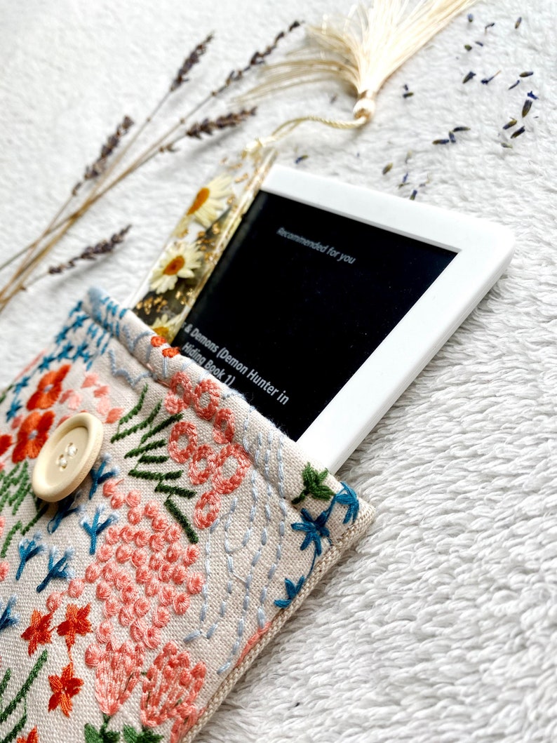 May include: A white e-reader with a floral bookmark in a beige fabric case with embroidered flowers. The e-reader screen displays the text "Recommended for you" and the title of a book, "A Demons Demon Hunter in Hiding Book 1".