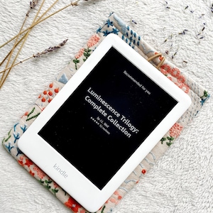 May include: A white Kindle e-reader with a black screen displaying the title "Luminescence Trilogy: Complete Collection" by J.L. Weil with a 5-star rating and 32 reviews. The Kindle is resting on a beige fabric case with floral embroidery.