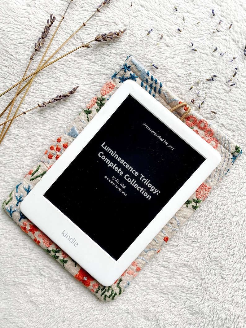 May include: A white Kindle e-reader with a black screen displaying the title "Luminescence Trilogy: Complete Collection" by J.L. Weil with a 5-star rating and 32 reviews. The Kindle is resting on a beige fabric case with floral embroidery.