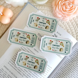 Puede incluir: Cuatro pegatinas en forma de billete de color azul verdoso y blanco con el texto "The Fantasy Book Club" y una ilustración caprichosa de un erizo, una ardilla y un pájaro. Las pegatinas están en un libro con texto en un idioma extranjero.