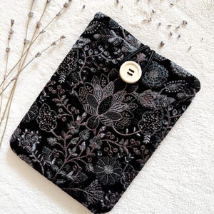 May include: Black embroidered book sleeve with a floral design. The sleeve has a button closure and a black elastic band. The embroidery features shades of grey and brown. The background is a white, textured surface.