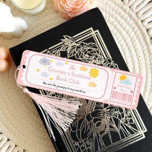 Puede incluir: Un marcap&aacute;ginas rectangular con dise&ntilde;o de entrada, borde rosa claro y borla rosa p&aacute;lido. Incluye ilustraciones de una nube gru&ntilde;ona y un sol sonriente, con el texto The Grumpy x Sunshine Book Club y You are the grumpy to my sunshine.