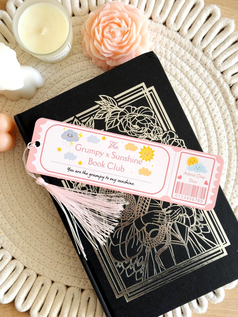Puede incluir: Un marcap&aacute;ginas rectangular con dise&ntilde;o de entrada, borde rosa claro y borla rosa p&aacute;lido. Incluye ilustraciones de una nube gru&ntilde;ona y un sol sonriente, con el texto The Grumpy x Sunshine Book Club y You are the grumpy to my sunshine.
