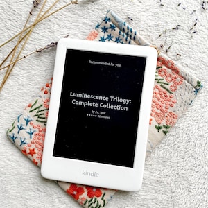 May include: A white Kindle e-reader with a black screen displaying the title "Luminescence Trilogy: Complete Collection" by J.L. Weil. The screen also shows a five-star rating and 32 reviews. The Kindle is resting on a beige fabric case with a floral embroidery design.