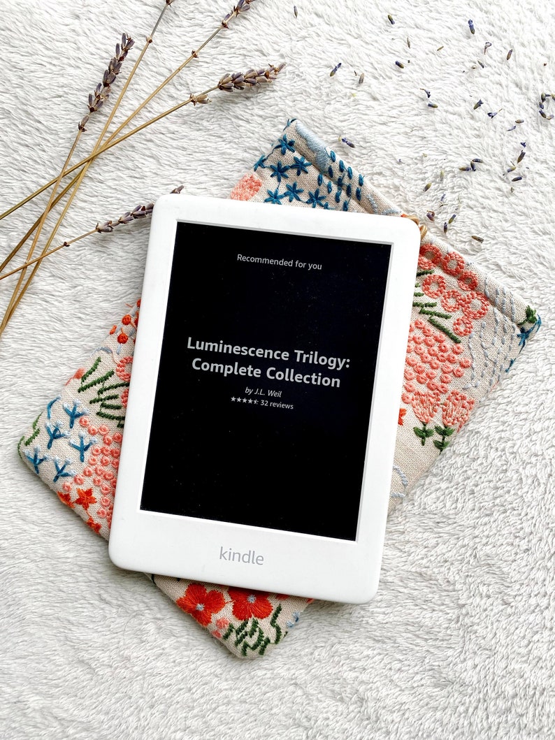 May include: A white Kindle e-reader with a black screen displaying the title "Luminescence Trilogy: Complete Collection" by J.L. Weil. The screen also shows a five-star rating and 32 reviews. The Kindle is resting on a beige fabric case with a floral embroidery design.