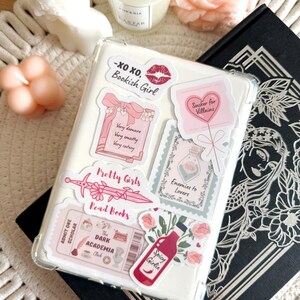 May include: A clear phone case with various stickers, including a pink heart with the text "Sucker for Villains", a pink and white sticker with the text "Pretty Girls Read Books", and a pink and white sticker with the text "The Dark Academia Club".