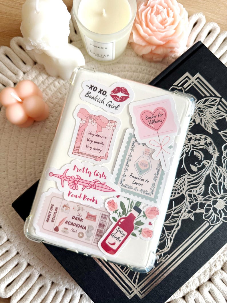 May include: A clear phone case with various stickers, including a pink heart with the text "Sucker for Villains", a pink and white sticker with the text "Pretty Girls Read Books", and a pink and white sticker with the text "The Dark Academia Club".