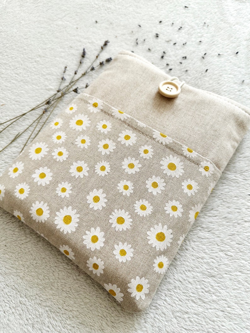 May include: A beige linen pouch with a white daisy pattern and a wooden button closure. The pouch is on a white fuzzy surface.