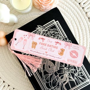 May include: A rectangular pink paper bookmark with a light pink tassel, featuring illustrations of champagne glasses, bows, teddy bears, and a red kiss mark. The text reads The Fake Dating Book Club, Admit One: Twisted Liar, and We're just playing a role, right? No feelings involved?