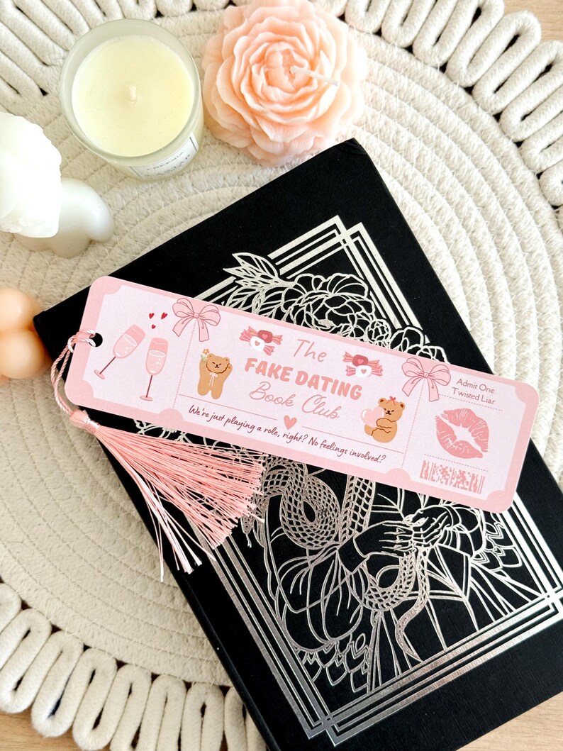 May include: A rectangular pink paper bookmark with a light pink tassel, featuring illustrations of champagne glasses, bows, teddy bears, and a red kiss mark. The text reads The Fake Dating Book Club, Admit One: Twisted Liar, and We're just playing a role, right? No feelings involved?