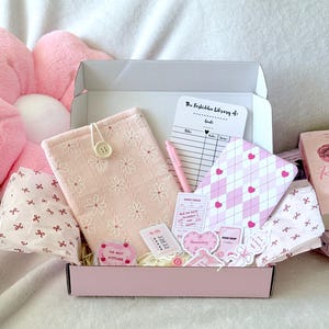 Op de afbeelding: Een roze en witte cadeaudoos gevuld met schrijfwaren en andere kleine items. De doos is versierd met een roze strik en de woorden "The Forbidden Library of Goals". De doos bevat een roze en wit notitieboekje, een roze pen, een roze en witte telefoonhoes, een roze en witte sticker en een roze en witte kaart.