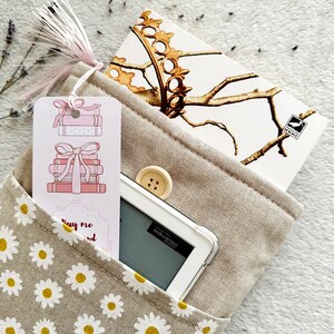 May include: A beige fabric book bag with a daisy print. The bag has a pocket on the front with a pink bookmark featuring a stack of books with a bow. A white e-reader and a notebook with a gold crown design are inside the bag.
