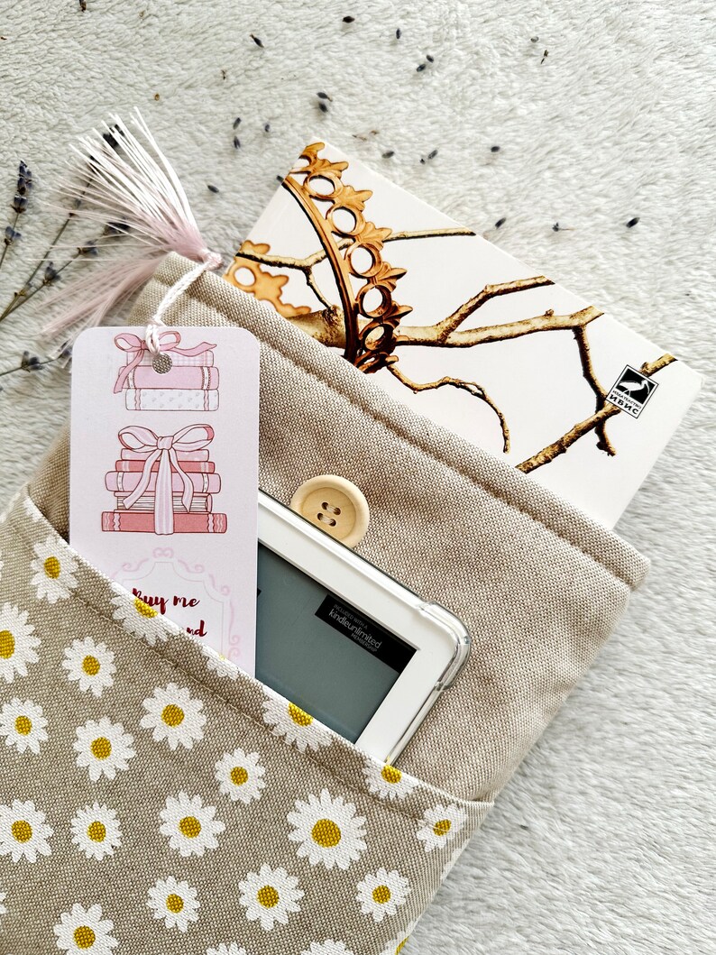 May include: A beige fabric book bag with a daisy print. The bag has a pocket on the front with a pink bookmark featuring a stack of books with a bow. A white e-reader and a notebook with a gold crown design are inside the bag.